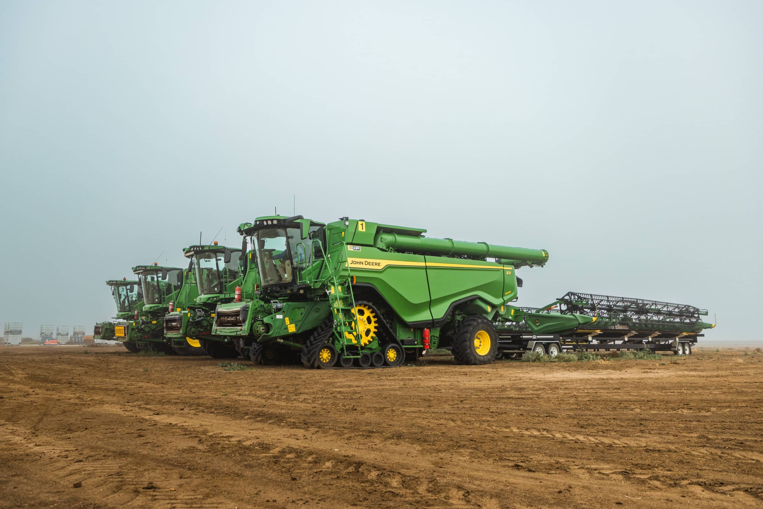 A line-up of green combine harvesters parked on bare earth under a hazy sky ahead of auction